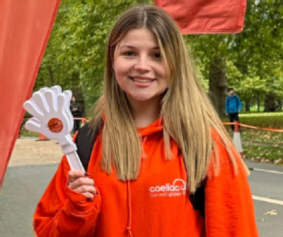 Coeliac UK fundraising coordinator Amy smiling with  wearing an orange Coeliac UK hoodie with a spring backdrop.